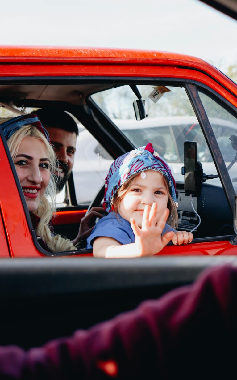 A cheerful family smiles and waves from inside a red car during a sunny road trip.